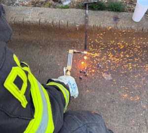 A person in firefighter gear uses an oxy-acetylene torch on a metal object at a curb, producing bright sparks that scatter across the concrete. A white plastic cup sits nearby on the curb.