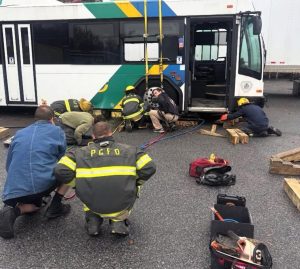 Five firefighters kneel and work together to rescue a mannequin trapped under a city bus during a training exercise. Tools, hoses, and wooden blocks are scattered on the ground near the bus.