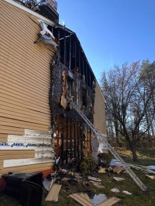 A house with severe fire damage along one exterior wall; siding is melted and burned, exposing charred wood and interior. A ladder leans against the building, and debris is scattered on the ground below.