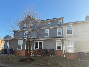 A gray and white multi-story townhouse with a bare tree in front, several air conditioning units along the brick foundation, and some debris scattered on the grass and sidewalk. The sky is clear and blue.