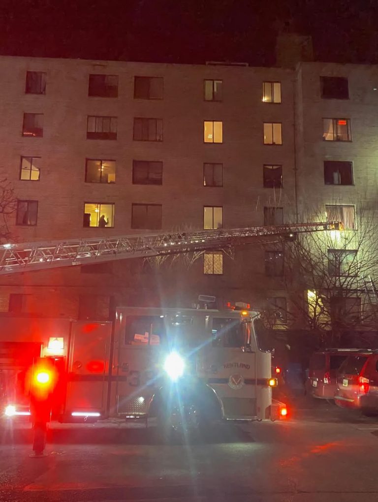 A fire truck with an extended ladder is parked outside a multi-story apartment building at night. Emergency lights are flashing, and some windows in the building are lit. Several cars are parked nearby.