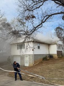 A firefighter unrolls a hose in front of a white house with smoke billowing from its windows, while another firefighter approaches the house. The yard is brown and leafless trees are visible.