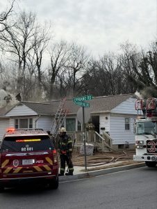 Firefighters respond to a house fire, with smoke coming from the roof. Emergency vehicles are parked outside, and ladders are propped against the house. Nearby, a street sign reads "Cipriano Rd" and "Old Barn Hill Ave.