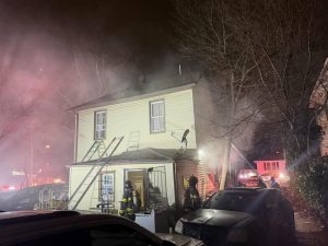 At night, firefighters stand outside a two-story house with smoke billowing from the roof. Ladders lean against the house, and emergency lights illuminate the scene. A car is parked in front, and bare trees surround the area.