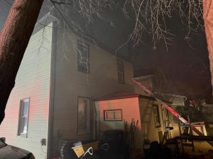 A two-story house at night with smoke and a fire truck ladder against the roof. Emergency lights cast a red glow on the siding. Outdoor equipment and tools are visible near the entrance. Leafless tree branches frame the scene.