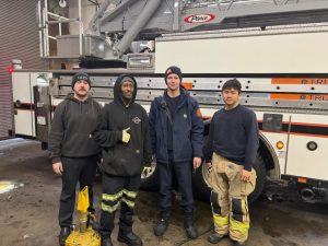 Four firefighters stand smiling in front of a fire truck inside a garage. Three wear dark uniforms, one wears tan fire-resistant pants, and equipment is visible around them.