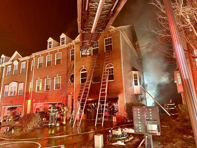 Firefighters respond to a nighttime fire at a brick townhouse, with smoke billowing from the building. Ladders extend to the upper floors, and fire hoses and equipment are spread across the wet ground.