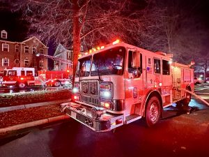 A fire truck with flashing lights is parked on a street at night. Another fire truck and a ladder reaching to a brick building are visible in the background, suggesting emergency response activity.