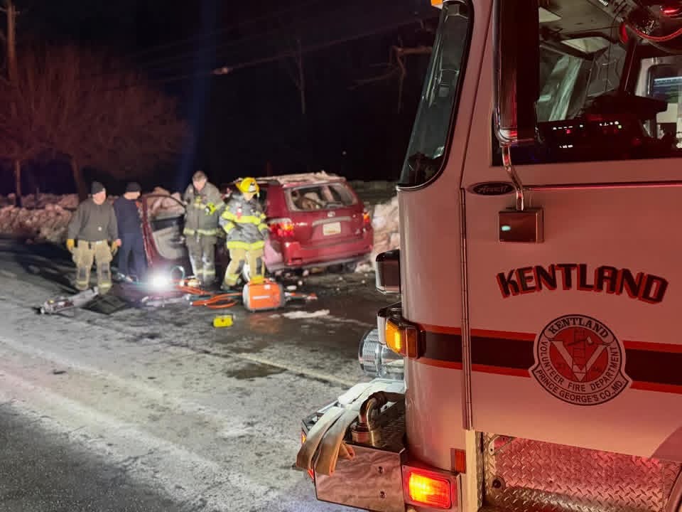 Firefighters and emergency responders work at night near a red vehicle involved in an accident on a snowy road, with fire department equipment and a Kentland fire truck visible in the foreground.