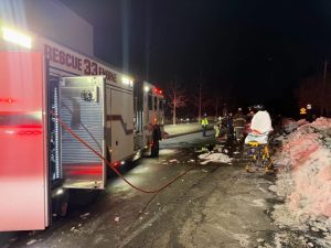 A fire truck with flashing lights and an extended hose is parked on a snowy, icy road at night. Emergency responders work nearby, and an empty stretcher is positioned on the roadside.