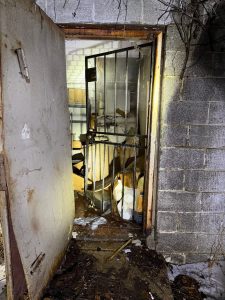 An open, rusty metal door leads to a cluttered room with shelves, boxes, and scattered debris. The floor is dirty, with snow and leaves outside the entrance. The scene is dimly lit, giving an abandoned feel.