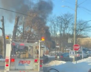 A fire truck with flashing lights is parked on a snowy street near a stop sign as thick black smoke rises from a fire in the background. Trees and several vehicles are visible nearby.