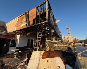 A burned building with charred debris and damaged exterior walls. Firefighters and ladders are present, and the ground is covered with scorched remains. The sky is clear and blue.