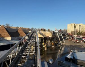 Firefighters work on the roof and ground near a heavily damaged building, using ladders and fire trucks. Water covers the pavement, and hoses are visible. Nearby, other buildings and an apartment complex are seen.