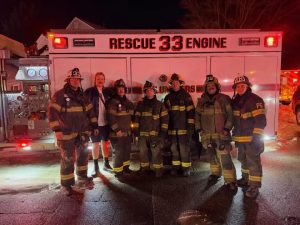 Six firefighters in full gear and one person in casual clothing stand together in front of a fire rescue truck labeled "Rescue 33 Engine" at night, illuminated by the vehicle’s lights.