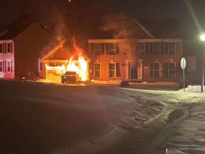 A house is on fire at night, with flames and smoke coming from the garage area. A vehicle in the driveway is also engulfed in flames. Snow covers the ground in front of the house.