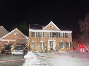 Firefighters work outside a two-story brick house at night, with fire hoses and ladders in use. The garage appears damaged, and a car is parked inside. Snow covers the ground, and emergency lights are visible on the right.