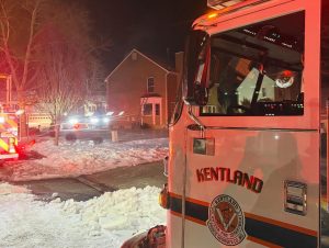 A Kentland fire truck is parked on a snowy residential street at night. Bright lights illuminate a house in the background as firefighters respond to an emergency.