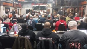 A group of people sit facing a table and a fire truck inside a firehouse, attending a community meeting. Several attendees wear jackets labeled "Brentwood" and "BVFD." Laptops and drinks are on the tables.