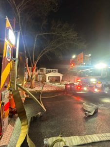Fire trucks and hoses are on the street at night in front of a house with smoke damage. Emergency lights illuminate the scene, and snow covers the ground near the sidewalk. A bare tree stands in the yard.