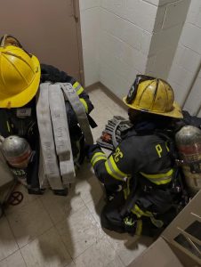 Two firefighters in full gear and yellow helmets are kneeling in a tiled hallway, handling fire hoses and equipment near a closed door.
