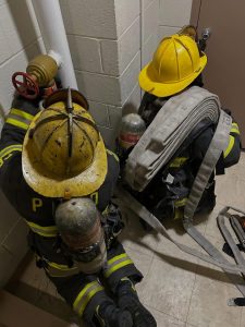 Two firefighters in full gear and yellow helmets kneel on a tiled floor near a wall, working with a fire hose and valve. One firefighter has a hose over their shoulder; both have oxygen tanks on their backs.