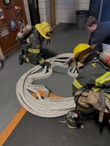 Two firefighters in full gear kneel on the floor, coiling a fire hose into a large, neat loop while another person watches nearby in a fire station. Fire helmets and equipment are visible.
