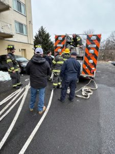 A group of firefighters and one other person stand near the back of a fire truck with orange and white stripes. Fire hoses are laid out on the wet ground, and one firefighter is on top of the truck.