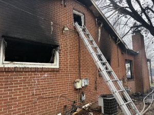 A brick house with heavy fire damage around the windows and roof. An aluminum ladder leans against the wall, leading to an open, charred second-story window. Debris is scattered near an outdoor AC unit.