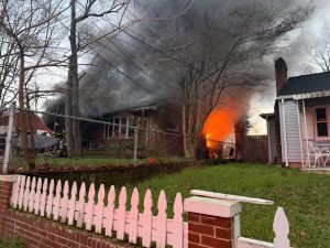 A house is on fire with large flames and thick black smoke rising; firefighters are visible near the burning home, while neighboring houses and a white picket fence are in the foreground.