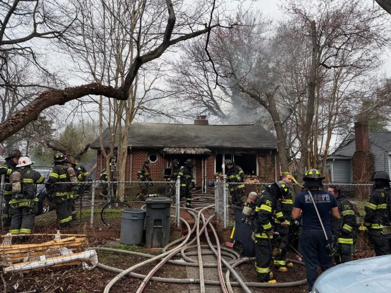 A group of firefighters in gear stand outside a small brick house with visible smoke damage and soot, working to control a fire. Hoses are spread across the yard, and trees surround the scene.