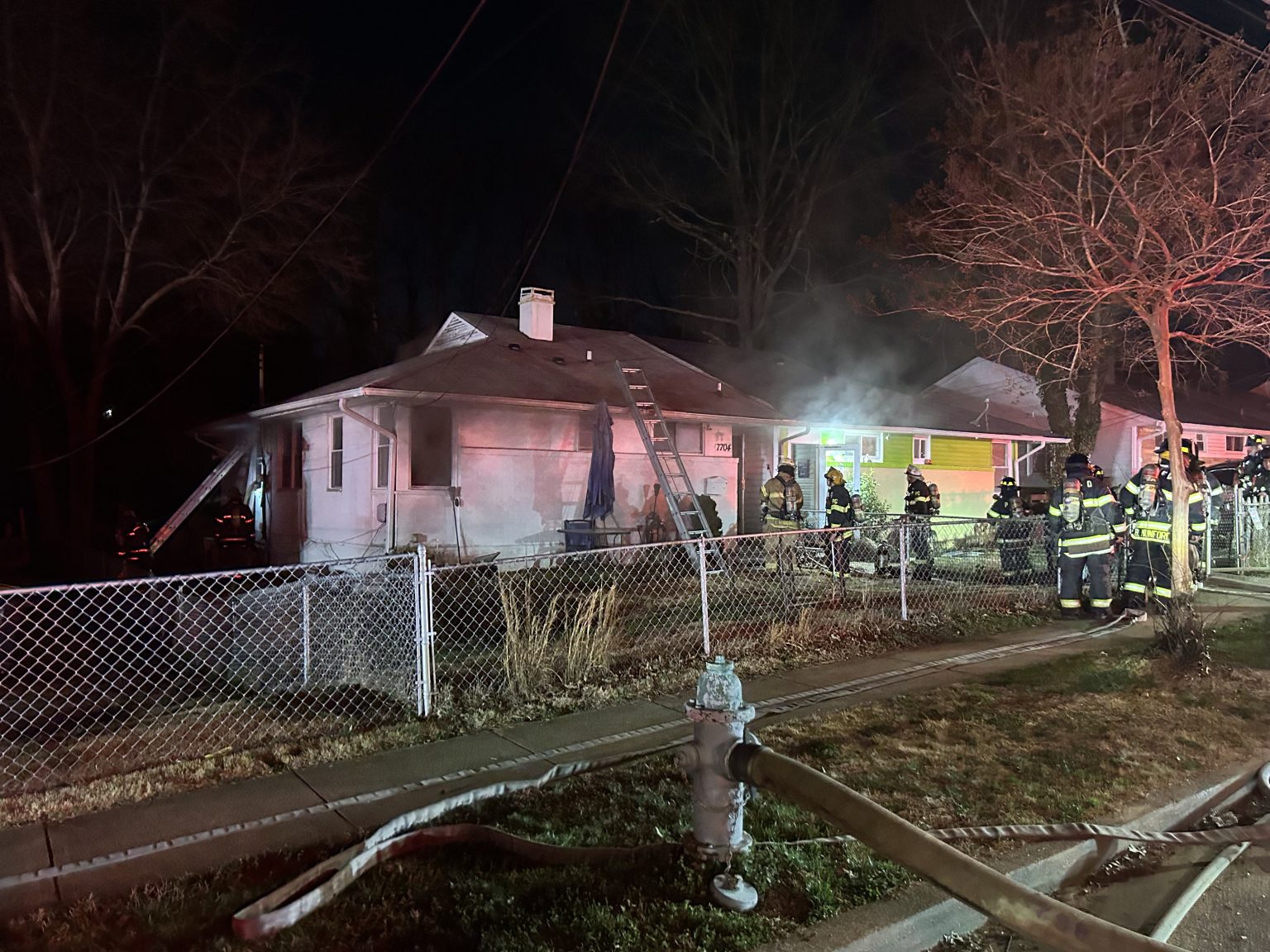 Firefighters respond to a house fire at night. Smoke rises from the white single-story home, with a ladder leaning against it. Fire hoses and a hydrant are visible, and several firefighters stand on the lawn and sidewalk.