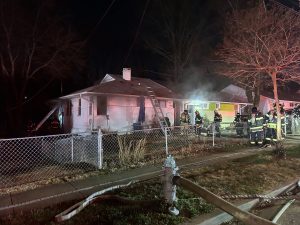 Firefighters respond to a house fire at night. Smoke rises from the white single-story home, with a ladder leaning against it. Fire hoses and a hydrant are visible, and several firefighters stand on the lawn and sidewalk.
