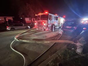 A fire truck with flashing lights is parked on a street at night, with hoses connected to a nearby fire hydrant. Several vehicles are parked along the curb, and houses are visible in the background.