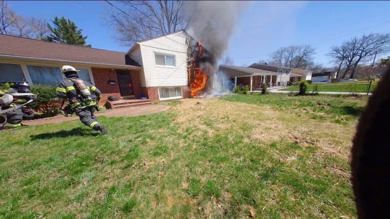 Firefighters approach a house with flames and smoke pouring from one side, while others stand nearby on the lawn. The sky is clear and the surrounding grass is green.