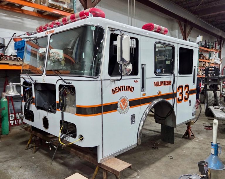 A white Kentland Volunteers fire truck with red lights and the number 33 is parked in a garage, undergoing repairs with missing front parts and exposed wires. Tools and equipment are visible in the background.