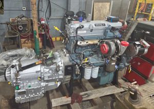 A large diesel engine and transmission assembly sit on wooden pallets in a workshop with tools, equipment, and machinery in the background. The engine appears newly rebuilt or clean, with various attached components visible.