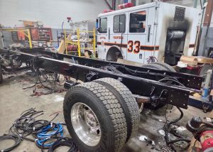 A fire truck with the cab marked "33 Volunteers" is being repaired in a workshop; its rear body is removed, exposing the chassis, wheels, wiring, and various tools and equipment scattered around.