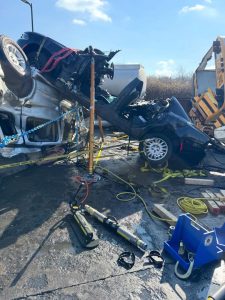 Two severely damaged cars are stacked on top of each other after a crash. Emergency rescue equipment, including hydraulic tools, cables, and caution tape, are visible around the scene. The area is sunny with blue skies.