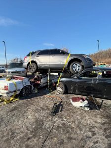 A silver SUV is strapped on top of two smashed cars, one white and one black, at a junkyard. Nearby, a van and other vehicles are visible, with a clear blue sky in the background.