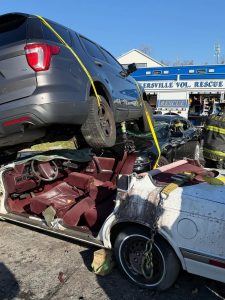 A dark SUV is perched on top of a white sedan with a crushed roof. Rescue workers and equipment are nearby. The background shows a blue rescue truck marked "RESCUE" and a building.