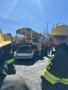Firefighters stand near a crash scene where a school bus is on top of a car. One firefighter works between the vehicles, while others watch. The sky is clear and blue in the background.