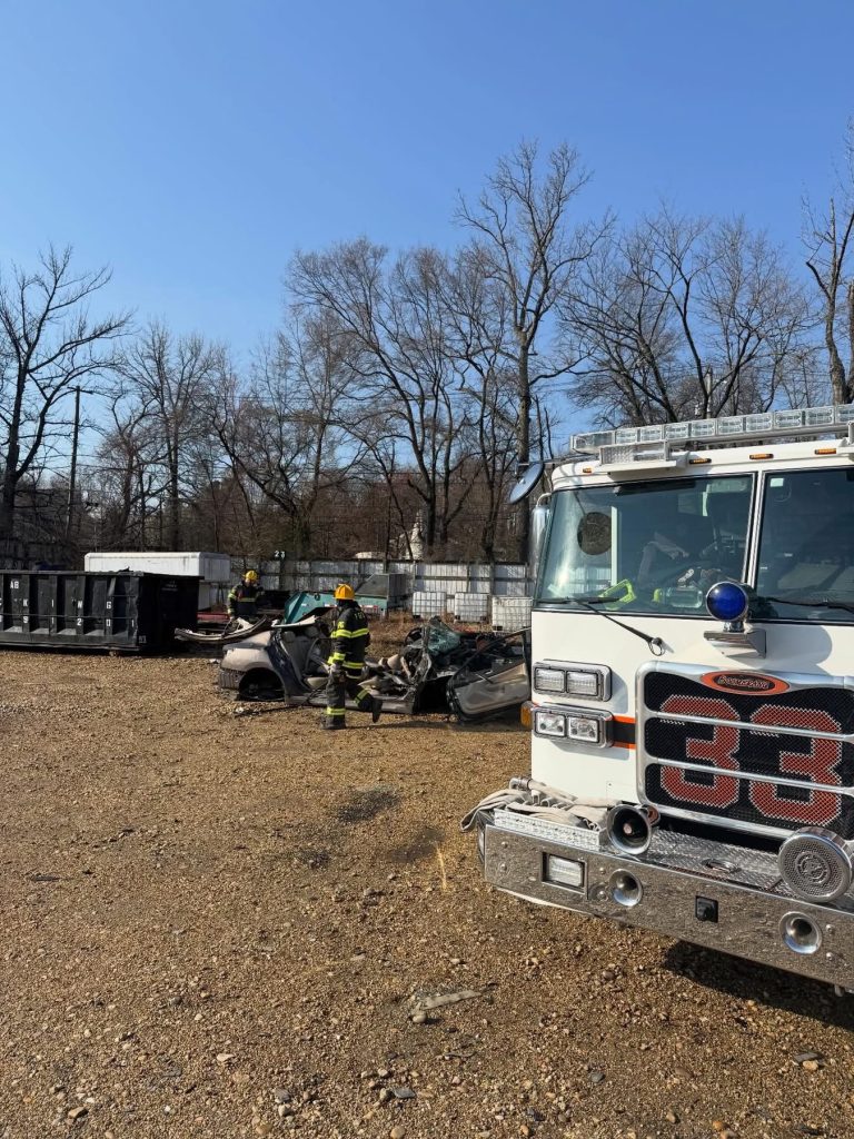 Firefighters in gear stand near a damaged car in a gravel lot with bare trees and a large fire truck labeled "33" in the foreground on a clear day.