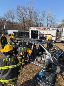 Firefighters in yellow helmets and PGFD uniforms use tools to cut apart a heavily damaged car during a rescue training exercise. A rescue vehicle and trees are visible in the background.