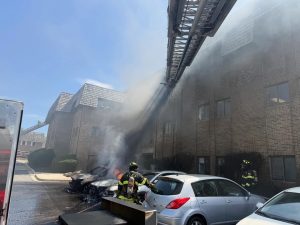 Firefighters extinguish a car fire in a parking lot next to a brick apartment building, with smoke rising and water spraying from a ladder truck above. Several cars are parked nearby, and flames are visible.