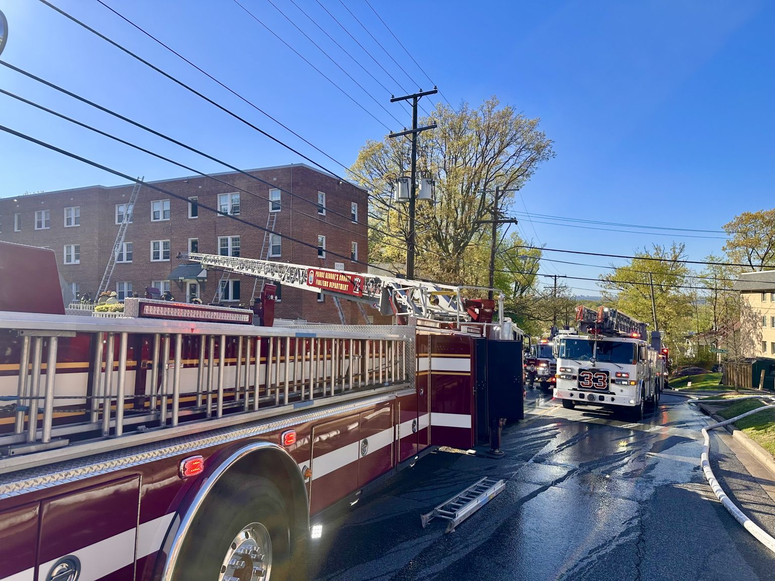 A fire truck with extended ladder is parked on a wet residential street near a brick apartment building, with another fire truck and emergency hoses visible in the background under a clear blue sky.