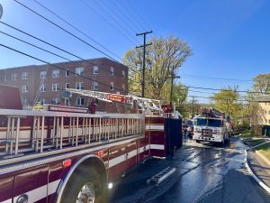 A fire truck with extended ladder is parked on a wet residential street near a brick apartment building, with another fire truck and emergency hoses visible in the background under a clear blue sky.