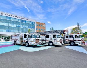 Three white fire trucks with "33" on the front are parked in front of the Wayne K. Curry Administration Building on a sunny day, with colorful circular patterns on the pavement.