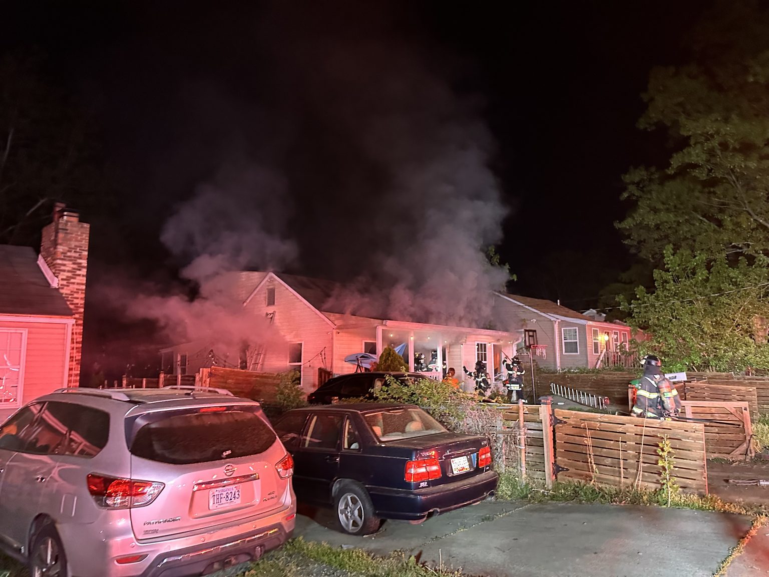 Smoke pours from a single-story house at night as firefighters work at the scene. Several cars are parked in the driveway and street; emergency lights illuminate the area.