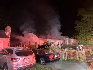 Smoke pours from a single-story house at night as firefighters work at the scene. Several cars are parked in the driveway and street; emergency lights illuminate the area.